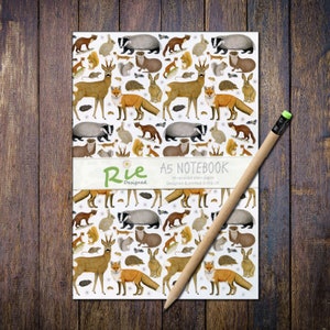 May include: A5 notebook featuring a repeating pattern of woodland creatures such as foxes, deer, and badgers. A pencil rests on the cover. The notebook has the text "Rie Designed" and "A5 Notebook" printed on it. 36 recycled plain pages.