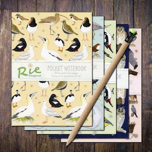 May include: A collection of pocket notebooks with bird designs and a pencil. The notebooks display different bird species on light yellow, blue, and pink backgrounds. The pencil is made of wood. The notebooks are labelled "Rie Designed POCKET NOTEBOOK" and "Designed & printed in the UK".