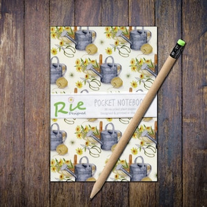 May include: Pocket notebook with a gardening theme, featuring a repeating pattern of watering cans, sunflowers, and twine. A pencil with a green eraser rests on the notebook. The notebook has the text "Rie Designed" and "Pocket Notebook".