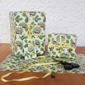 May include: Two wrapped gifts with a botanical print featuring green leaves, red berries, and black berries on a white background. The gifts are tied with yellow ribbon and a matching gift tag. Scissors and wrapping paper are also visible.
