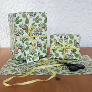 May include: Two wrapped gifts with botanical-themed wrapping paper featuring green leaves, red berries, and yellow accents. The gifts are tied with yellow ribbon and a matching gift tag. A pair of scissors and wrapping paper are also visible.