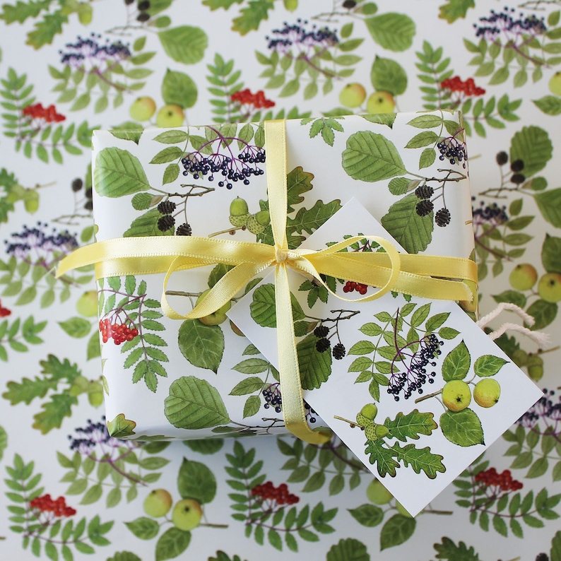 May include: A wrapped gift with a white background featuring a botanical pattern of green leaves, red berries, and green apples. The gift is tied with a yellow ribbon and a matching tag.