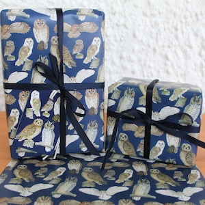 May include: Two wrapped gifts and a sheet of wrapping paper featuring a navy blue background with various owl illustrations in shades of brown, white, and tan. The gifts are tied with dark blue ribbons. The owls are depicted in different poses.
