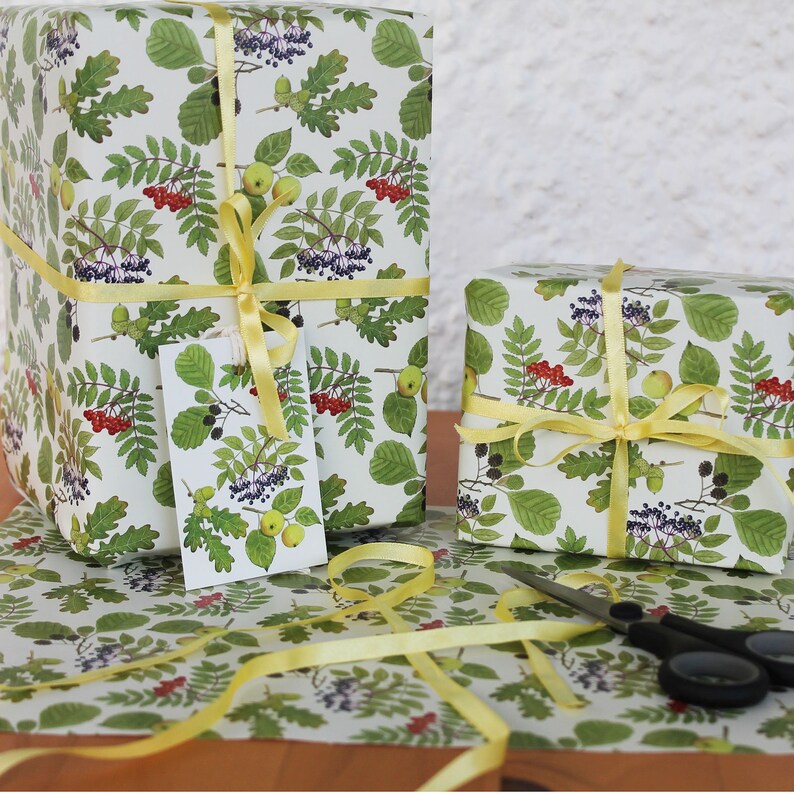 May include: Two wrapped gifts with botanical-themed wrapping paper featuring green leaves, red berries, and yellow apples. The gifts are tied with yellow ribbon. A gift tag with the same design is attached to one gift. Scissors and wrapping paper are also visible.