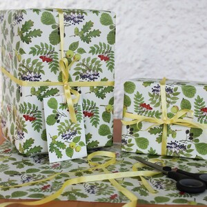 May include: Two wrapped gifts with botanical-themed wrapping paper featuring green leaves, red berries, and yellow ribbons. A matching gift tag is attached to one package. Scissors and extra ribbon are also visible.
