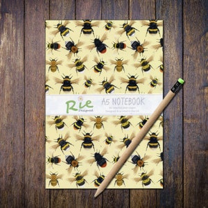 May include: A5 notebook featuring a repeating pattern of black and yellow bumblebees on a yellow background. The notebook has a white band with the text "Rie Designed" and "A5 NOTEBOOK". A wooden pencil is placed on the notebook.