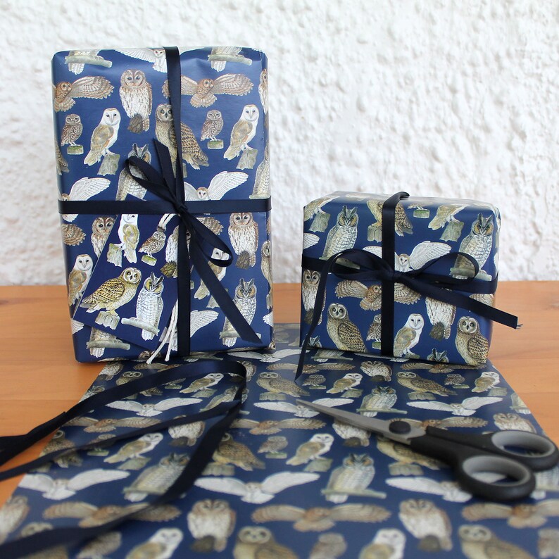 May include: Two wrapped gifts with navy blue owl-patterned paper and black ribbon. A matching tag is attached to one gift. A roll of the same paper and scissors are on a wooden surface. The owl illustrations are in shades of brown and white.