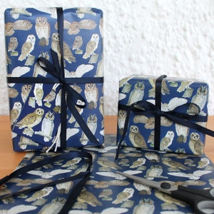 May include: Two wrapped gifts and wrapping paper featuring a navy blue background with various owl illustrations in shades of brown, white, and tan. The gifts are tied with a dark blue ribbon. A gift tag with an owl illustration is attached to one gift.