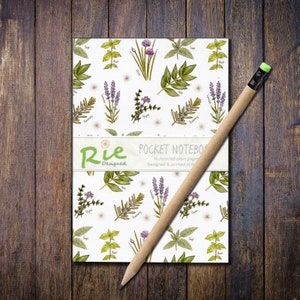 May include: A pocket notebook with a white cover featuring botanical illustrations of herbs. A brown pencil with a green eraser rests diagonally across the notebook, which is on a wooden surface. The notebook has 36 recycled plain pages.