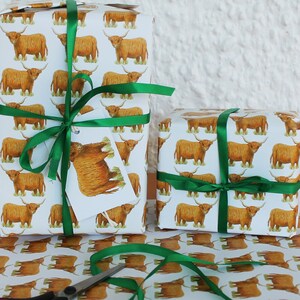May include: Two wrapped gifts with white paper featuring a repeating pattern of brown Highland cows. The gifts are tied with green satin ribbon and a tag with a cow illustration. Scissors and more wrapping paper are visible.