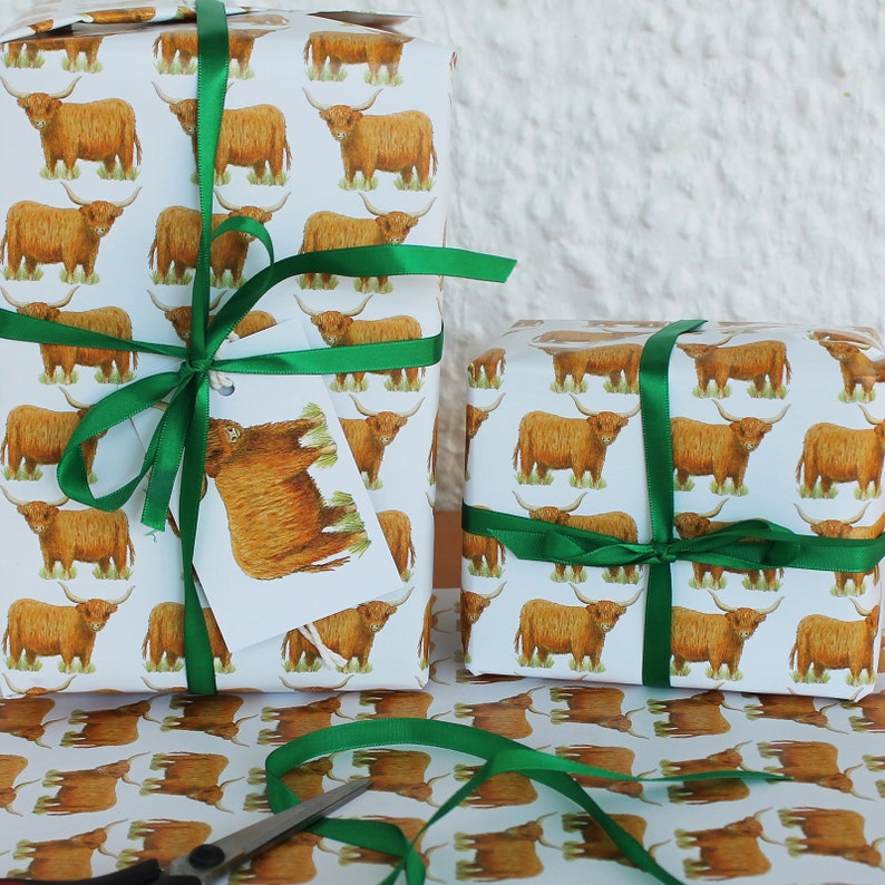 May include: Two wrapped gifts with white paper featuring a repeating pattern of brown Highland cows. The gifts are tied with green satin ribbon and a tag with a cow illustration. Scissors and more wrapping paper are visible.