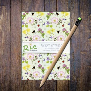 May include: A pocket notebook with a floral design of pink roses, yellow flowers, purple bellflowers, ladybirds, and bees. The notebook has a white band with the text "Rie Designed POCKET NOTEBOOK" and a pencil with a green eraser.