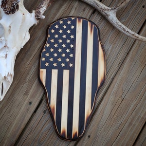 May include: A wooden deer skull plaque with an American flag design burned into the wood. The flag is black and white with a distressed look. The plaque is on a wooden surface.