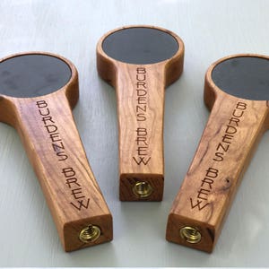 Puede incluir: Tres tiradores de cerveza de madera con tapas de pizarra negra. Cada tirador est&aacute; grabado con el texto "Burdens Brew".