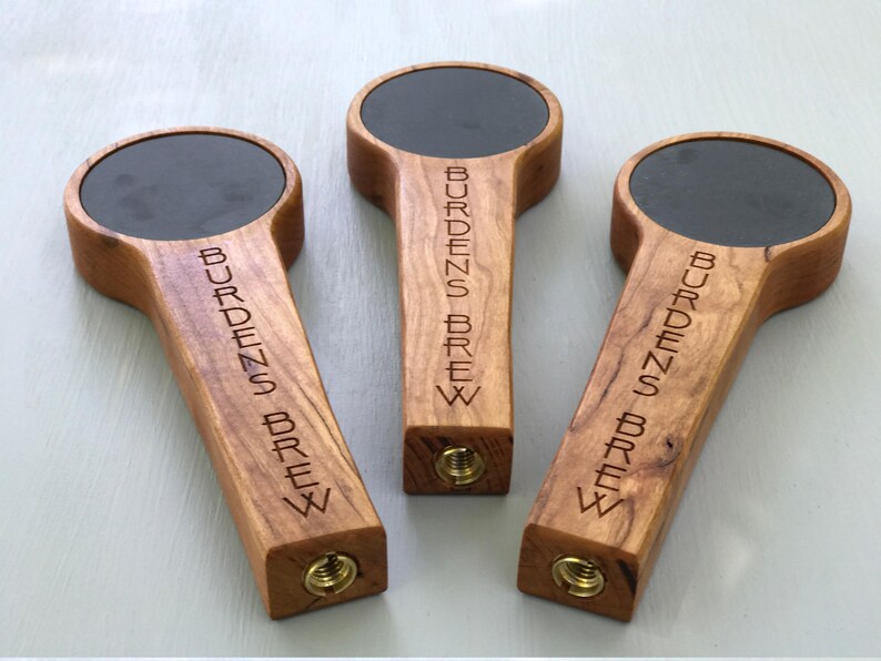 Puede incluir: Tres tiradores de cerveza de madera con tapas de pizarra negra. Cada tirador est&aacute; grabado con el texto "Burdens Brew".