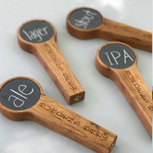Puede incluir: Cuatro tiradores de cerveza de madera con letreros de pizarra. Los letreros est&aacute;n etiquetados como "lager", "stout", "IPA" y "ale".