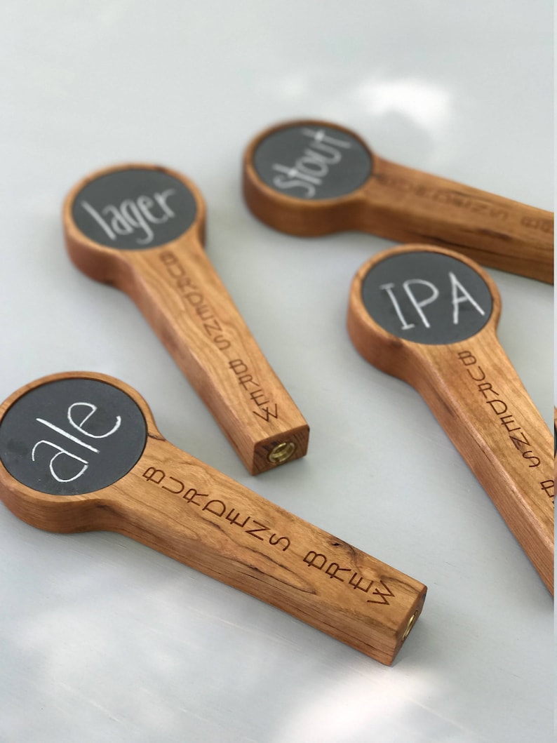 Puede incluir: Cuatro tiradores de cerveza de madera con letreros de pizarra. Los letreros est&aacute;n etiquetados como "lager", "stout", "IPA" y "ale".