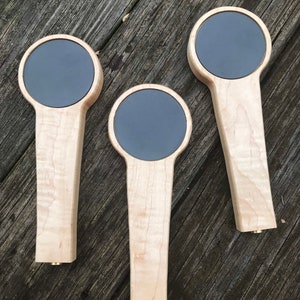 Puede incluir: Tres tiradores de cerveza de madera con insertos circulares negros. Los tiradores est&aacute;n hechos de madera de arce con un acabado natural.