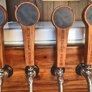 Puede incluir: Cuatro tiradores de cerveza de madera con tapas de pizarra negra. Cada tirador est&aacute; grabado con el texto "Burdens Brew".