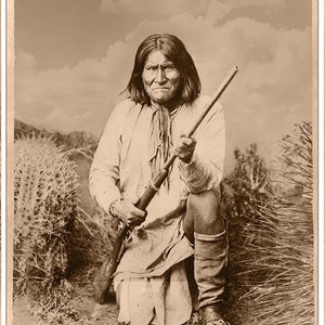 Op de afbeelding: Een zwart-witfoto van Geronimo, een Apache-leider, knielend met een geweer in zijn handen. Hij draagt een shirt met lange mouwen en een broek. De foto is buiten gemaakt en toont een grasachtige achtergrond.