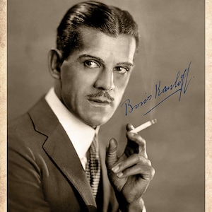 Puede incluir: Un retrato en blanco y negro de un hombre con traje, bigote y sosteniendo un cigarrillo. El hombre mira directamente a la cámara. La imagen está firmada "Boris Karloff".