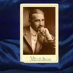 May include: A black and white photograph of L. Frank Baum, the author of The Wonderful Wizard of Oz. The photo shows Baum with a moustache and glasses, looking thoughtfully off to the side. He is wearing a suit and tie.