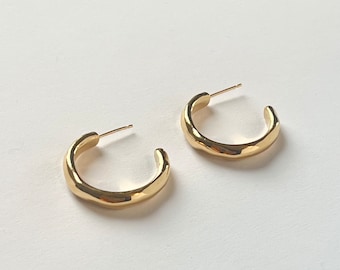 May include: A pair of gold-colored hoop earrings. The earrings have a thick, rounded design with a slightly textured surface. The posts are thin and extend from the top of the hoops. The earrings are displayed on a white background.