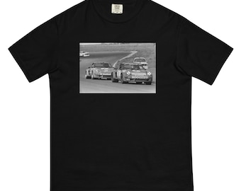 May include: Black and white photograph of two vintage race cars on a track. The cars are both Porsche 911s, one with the number 36 and the other with the number 90. The cars are in motion and the photo is taken from a low angle.