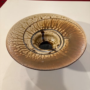 May include: A ceramic bowl with a unique design. The bowl features concentric circles and a central well. The exterior has a textured, organic pattern in shades of brown and tan. The interior of the well is dark.
