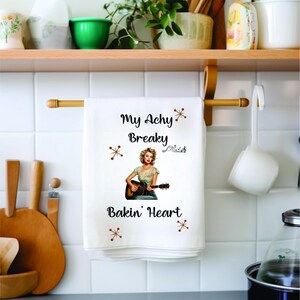Puede incluir: Toalla de cocina blanca con una ilustración retro de una mujer tocando la guitarra. El texto en la toalla dice "My Achy Breaky Bakin' Heart" con estrellas marrones.