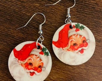 May include: A pair of round, white, wooden earrings featuring a vintage image of Santa Claus's face. The earrings have silver hooks.