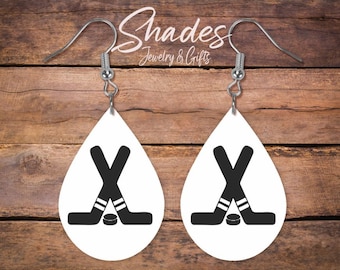 May include: White teardrop earrings featuring a black hockey stick and puck design. The earrings have silver-colored hooks. The text "Shades Jewelry & Gifts" is in the background.