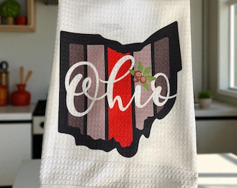 May include: White kitchen towel featuring the state of Ohio in a striped design with the word "Ohio" in a cursive font. The stripes are in shades of brown, red, and gray. A small floral accent is on the right side of the word.