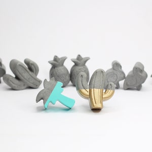 Puede incluir: Un conjunto de nueve pequeñas figuritas de hormigón en varios temas tropicales y desérticos. Las figuritas están pintadas en tonos de gris, turquesa y dorado. Las figuritas incluyen una palmera, un cactus, una piña y un flamenco.