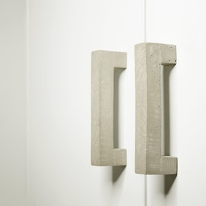 Może przedstawiać: Dwa betonowe uchwyty do drzwi szafki w jasnoszarym kolorze. Uchwyty mają kształt prostokątny z wycięciem w środku na uchwyt.
