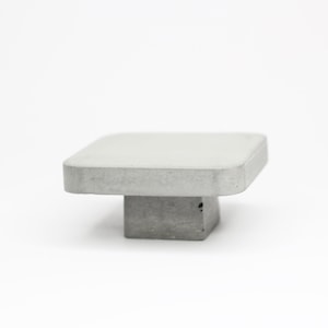Könnte beinhalten: Ein quadratischer Betonknauf mit abgerundeter Oberseite und quadratischem Sockel. Der Knauf ist hellgrau.