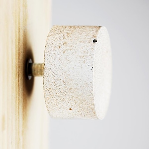 Peut inclure: Bouton en béton peint en blanc avec une texture rugueuse et un petit trou au centre. Le bouton est fixé à une surface en bois.