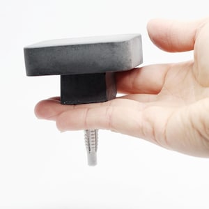 May include: A gray concrete square top with a black base and a silver screw. The top is held in a hand.