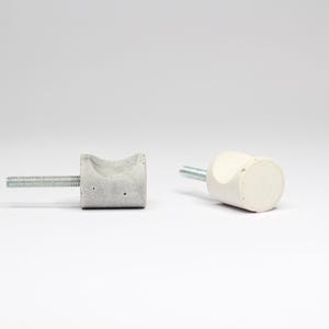 Pode incluir: Dois botões de armário, um é de concreto cinza e o outro é branco. Ambos têm um parafuso para instalação.