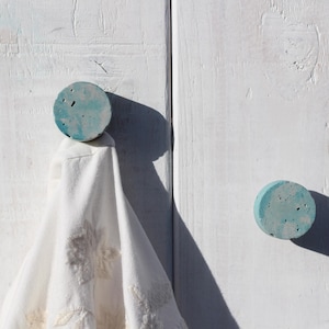 May include: Two round, blue-green concrete wall hooks fixed to a white wooden surface. One hook supports a white cloth with floral embroidery. The hooks are circular and have a speckled texture.