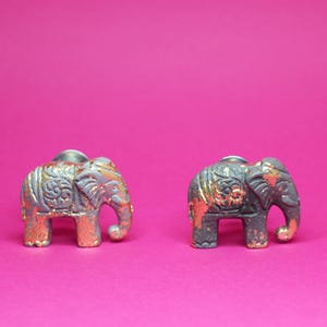 Könnte beinhalten: Zwei dekorative Elefantenfiguren mit aufwendigen Details und metallischem Finish. Die Elefanten sind grau und goldfarben bemalt, mit einem Hauch von Orange, vor einem leuchtend pinkfarbenen Hintergrund. Diese kleinen Skulpturen werden wahrscheinlich als Schrankknöpfe verwendet.