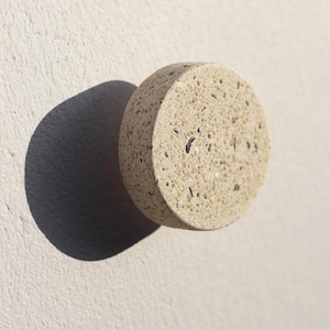 Op de afbeelding: Een ronde, beige betonnen knop met een gespikkelde textuur. De knop werpt een donkere schaduw op een lichtbeige muur. De knop is een decoratief element, mogelijk voor een kast of lade.
