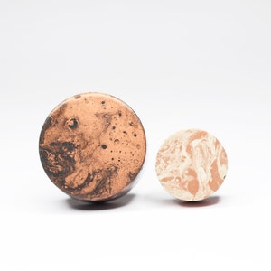 May include: Two round coasters with a marbled design. The larger coaster is brown with black marbling, and the smaller coaster is white with light brown marbling.