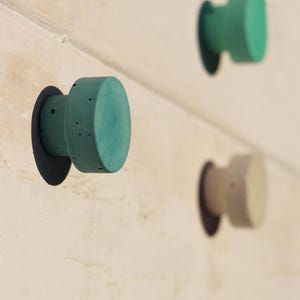 Può includere: Primo piano di tre pomelli per armadi in cemento nei toni del verde acqua e beige. Ogni pomello ha una faccia circolare e un corpo cilindrico, montato su una base circolare grigio scuro. I pomelli sono attaccati a una superficie chiara.
