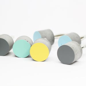 Puede incluir: Una colección de nueve pomos redondos de hormigón con tapas de diferentes colores. Los colores incluyen blanco, gris, amarillo, azul claro y turquesa.
