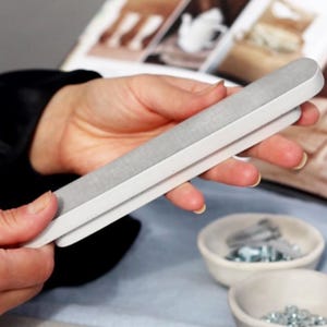 May include: A white nail file with a gray surface. The file is held in a person's hand.