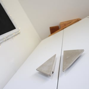 May include: Two triangular concrete cabinet door pulls in a light grey colour. The pulls are mounted on a white cabinet door.