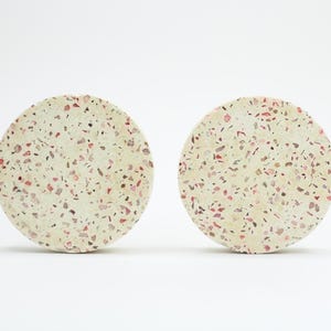 Può includere: Due sottobicchieri rotondi con un design maculato. I sottobicchieri sono beige con macchie rosse e nere. Sono realizzati in un materiale solido e adatti per appoggiare bevande.