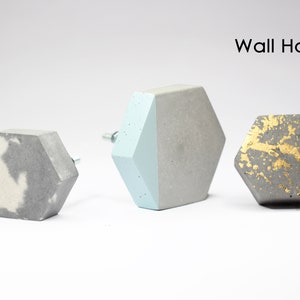 Könnte beinhalten: Drei sechseckige Wandhaken aus Beton. Die Haken sind in verschiedenen Farben: grau mit weißen Akzenten, hellblau und grau sowie grau mit goldenen Akzenten.