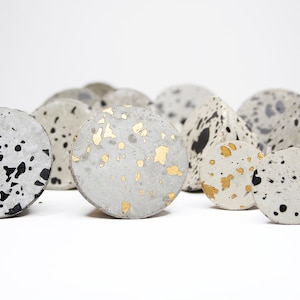 Peut inclure: Une collection de boutons en béton ronds et coniques avec des mouchetures noires et dorées. Les boutons sont en béton gris clair et ont une finition mouchetée.